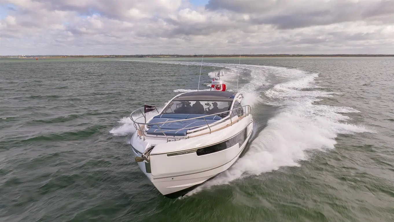 A sleek motorboat with a flag glides through calm waters, leaving a wake, against a backdrop of cloudy skies.